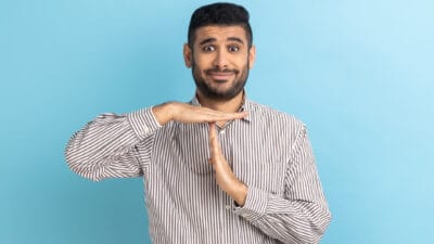 A man with a heavy facial hair growth and a comical look on his face holds his hands in a 'time out' gesture.