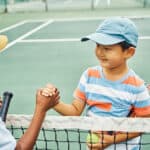 Two young boys with tennis racquets and wearing caps shake hands over a tennis ten on a tennie court.