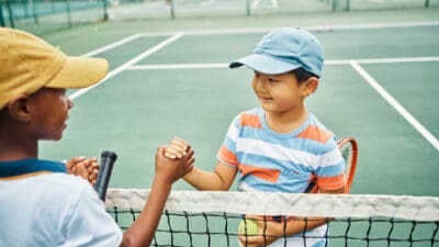 Two young boys with tennis racquets and wearing caps shake hands over a tennis ten on a tennie court.