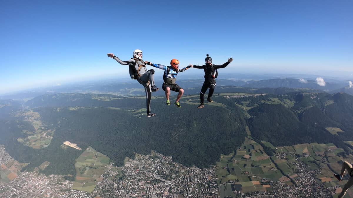 Three sky divers link arms in mid-air high above the earth below with a green panorama below them against a clear blue sky.
