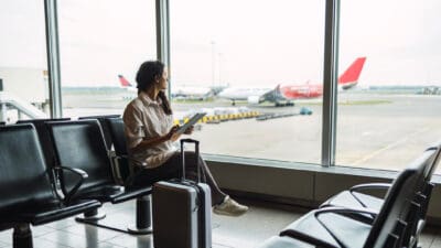 Woman on a tablet waiting in for her flight in an airport and looking through a window.