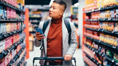 A man pushes a supermarket trolley with phone in hand down a supermarket aisle looking at the products on the shelves.