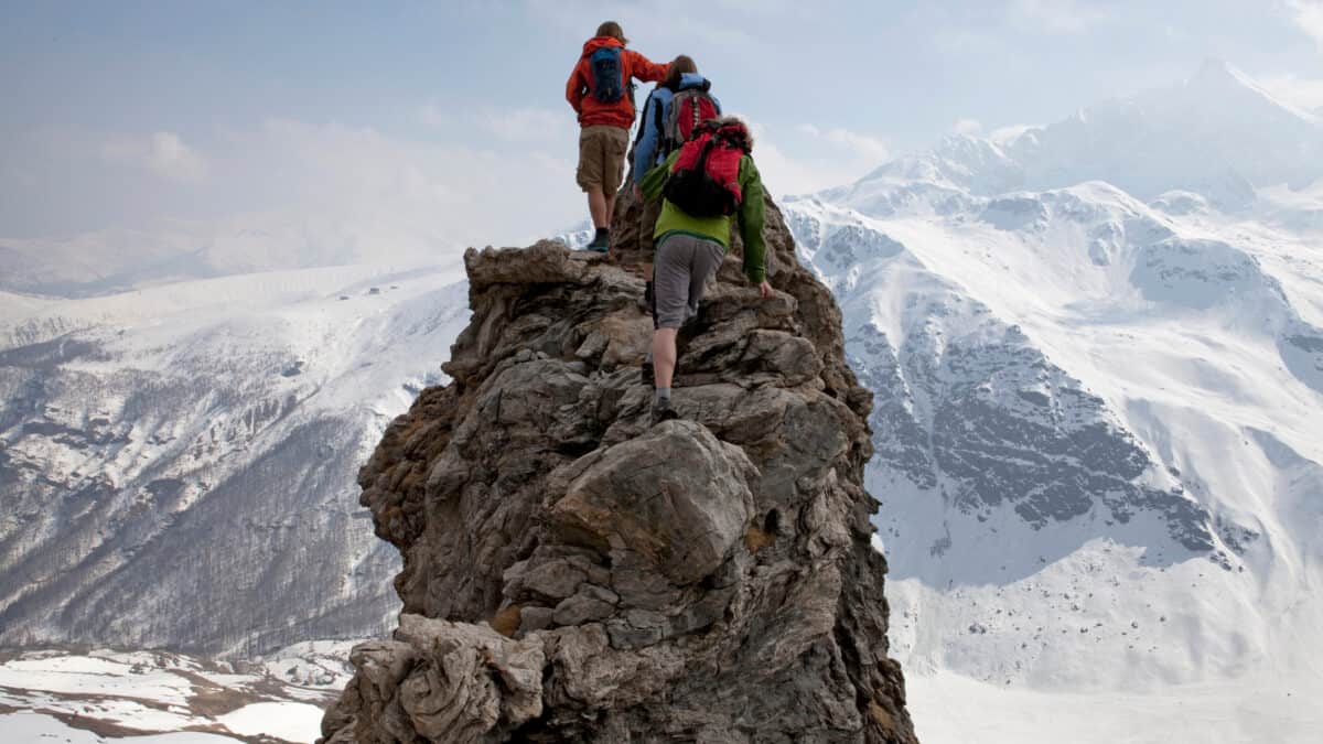 Three climbers scramble up a rocky peak overlooking a vast snow covered mountain range with an icy blue sky beyond them.