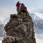 Three climbers scramble up a rocky peak overlooking a vast snow covered mountain range with an icy blue sky beyond them.