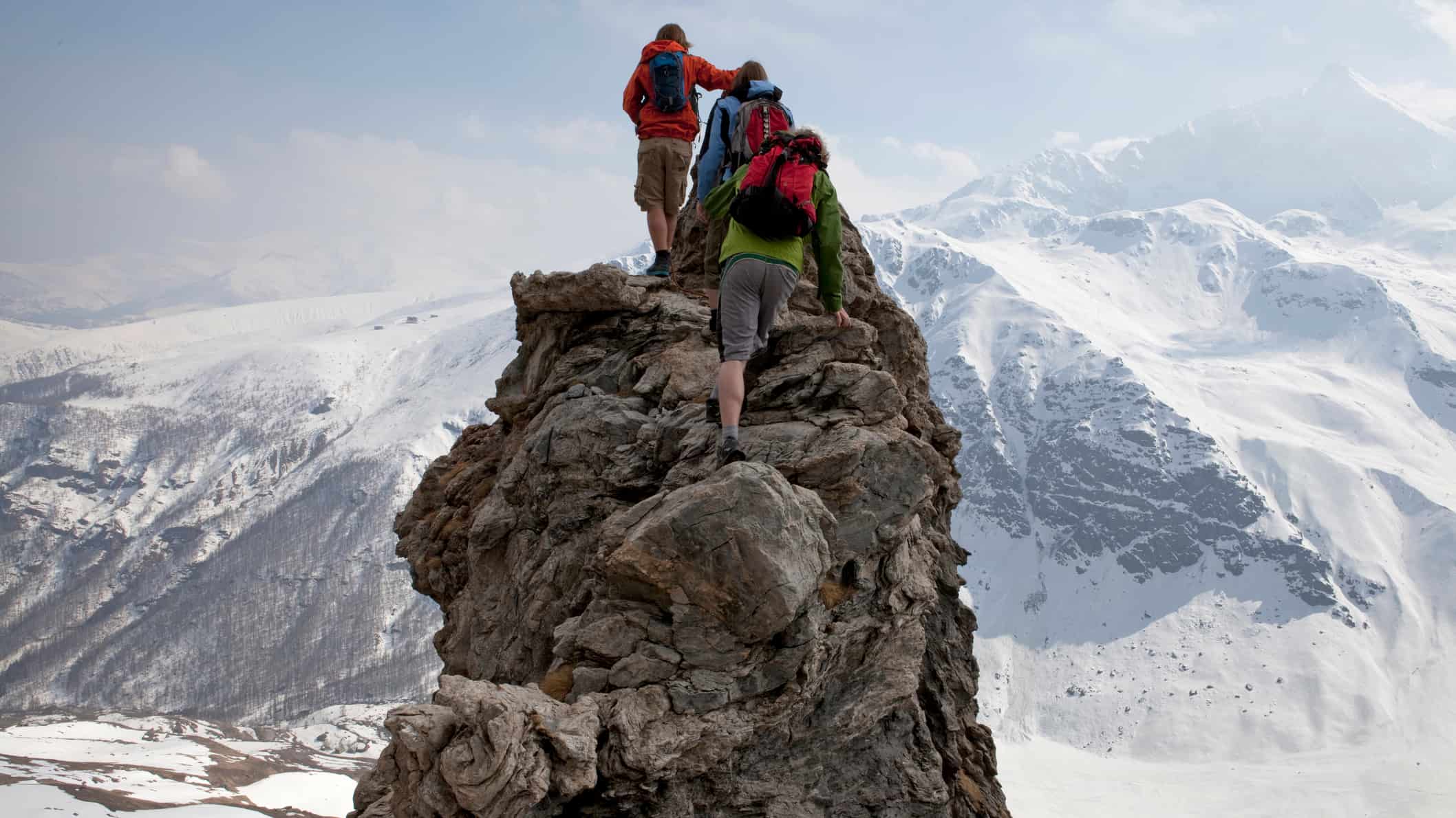 Three climbers scramble up a rocky peak overlooking a vast snow covered mountain range with an icy blue sky beyond them.