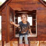A cute young girl wearing gumboots and play clothes holds open the door of her wooden cubby house as she sits and smiles in a backyard outdoor setting.