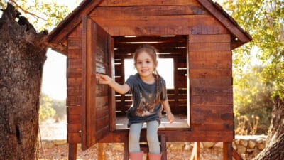 A cute young girl wearing gumboots and play clothes holds open the door of her wooden cubby house as she sits and smiles in a backyard outdoor setting.
