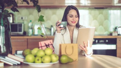 Woman with a coffee mug in one hand and a tablet in another along with pears on the table, symbolising inflation.