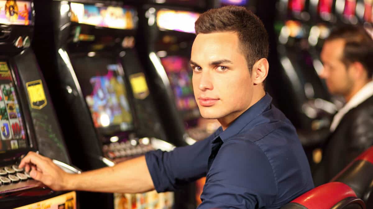A young man sits at a poker machine with a serious look on his face in a casino or club setting.