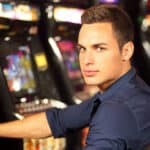 A young man sits at a poker machine with a serious look on his face in a casino or club setting.