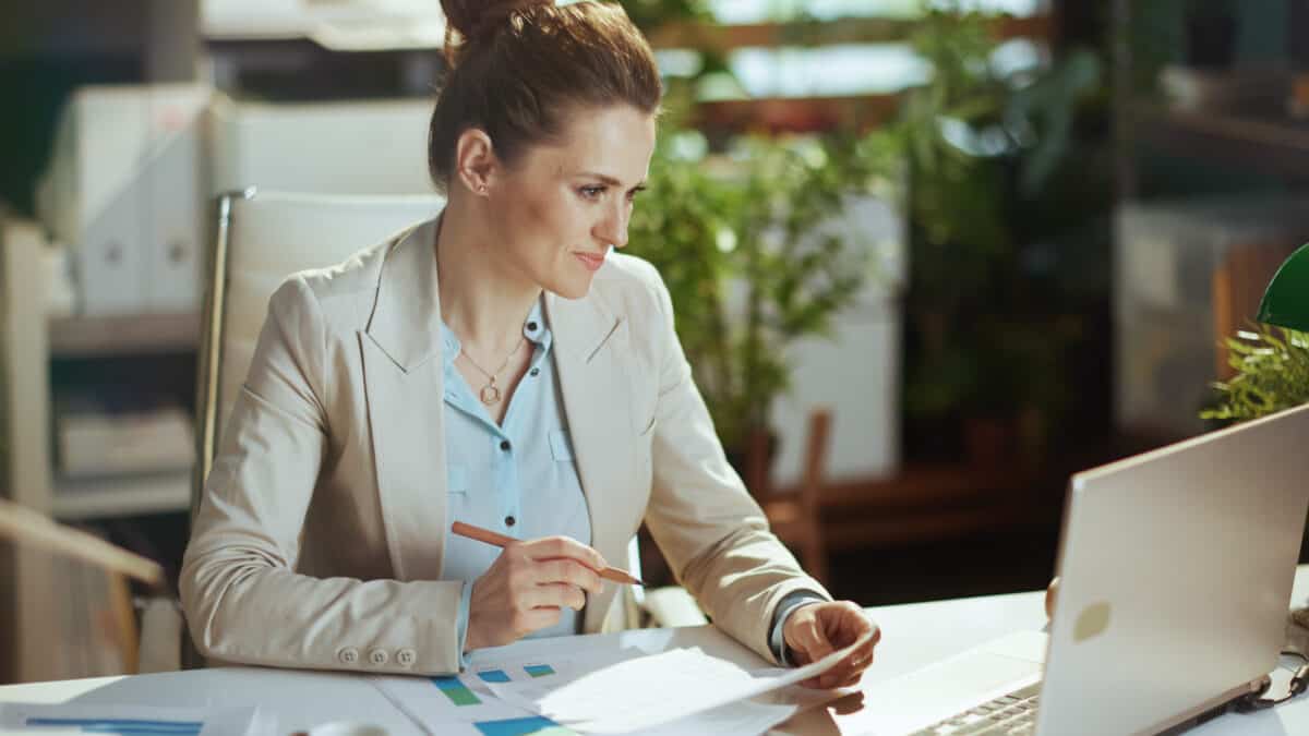 Modern accountant woman in a light business suit in modern green office with documents and laptop.