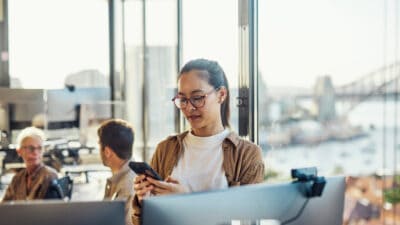 A woman stands at her desk looking a her phone with a panoramic view of the harbour bridge in the windows behind her with work colleagues in the background.