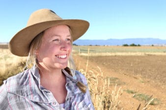 An Australian farming woman of the land wears an akubra hat and work shirt smiles broadly as she looks out over turned soil paddocks with a mountain range far off in the distance and blue sky above.