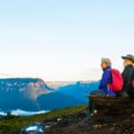 An older couple of retirement age and wearing hiking gear sit on a rocky outcrop gazing out at a sensational view of a rock formation and a waterway in the Australian bush.