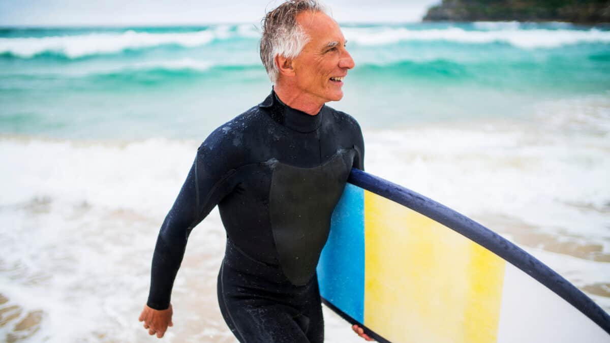 A man in his late 60s, retirement age, emerges from the Australian surf carrying a surfboard under his arm and wearing a wetsuit.