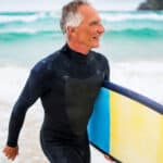 A man in his late 60s, retirement age, emerges from the Australian surf carrying a surfboard under his arm and wearing a wetsuit.