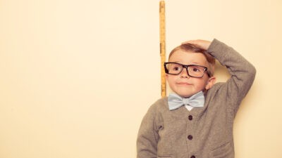 A cute little boy, short in height, wearing glasses, old-fashioned bow tie and cardigan stands against a wall near a tape measure with his hand at the top of his head as though to measure his height.