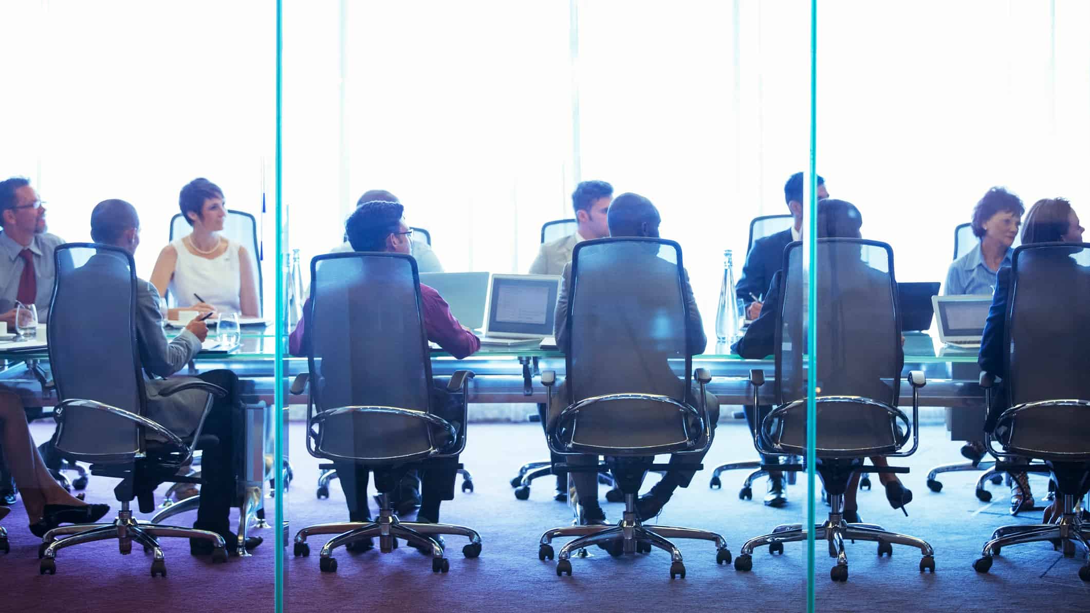 A view through a glass wall into a board room where people are sitting in chairs around a long table, some with their backs to the front of the picture, others racing the front.