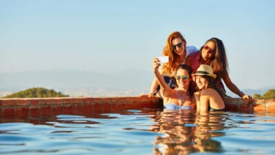 Happy female friends taking self portrait through mobile phone at pool's edge, symbolising passive income.