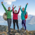 Three hikers lift their arms in jubilation as they reach a rocky peak overlooking a sensational view of water and mountains with a blue sky surrounding them.