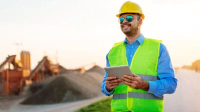 Image of young successful engineer, with blueprints, notepad and digital tablet, observing the project implementation on construction site and in mine.