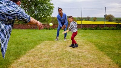 Kid swinging his bat and playing backyard cricket with his parents.