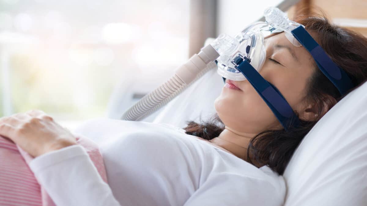 Senior woman using cpap machine to stop choking and snoring from obstructive sleep apnoea with bokeh and morning light background.