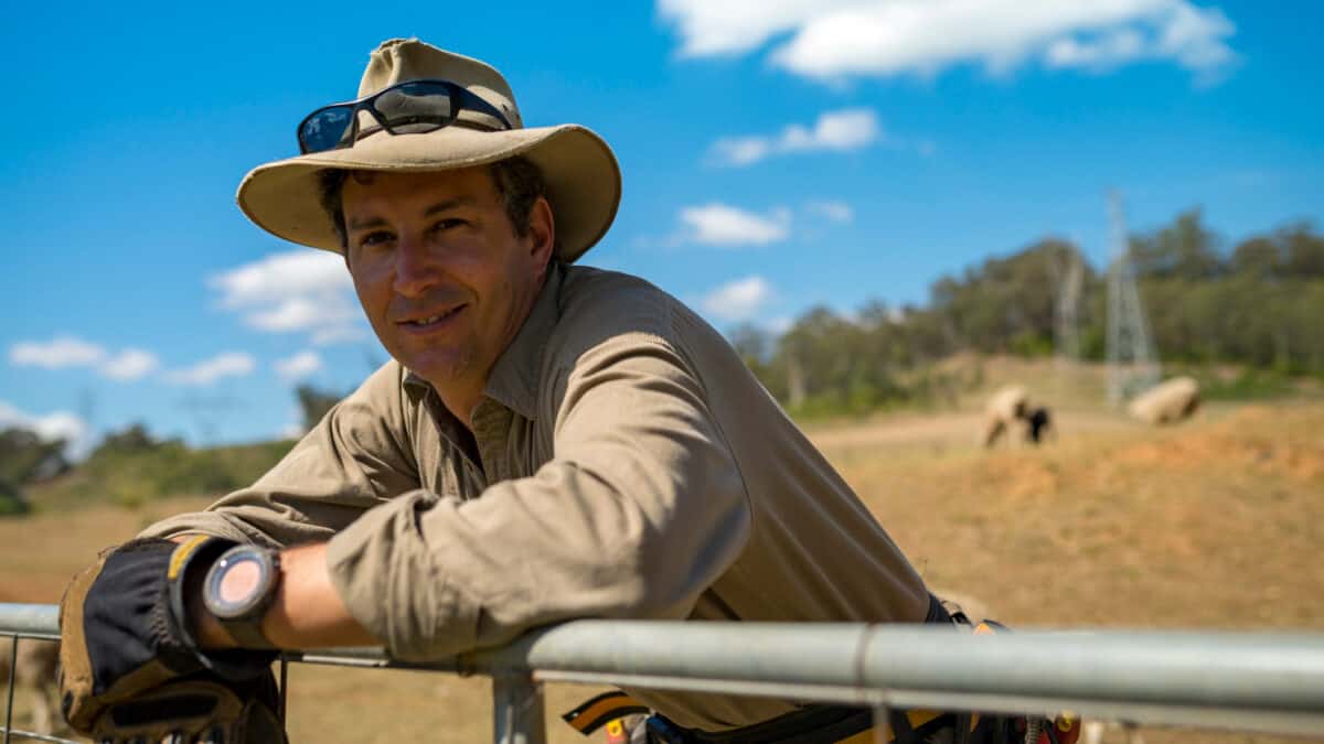 An Australian farmer wearing a beaten-up akubra hat and work shirt leans on a fence with livestock in the background and a blue sky above.
