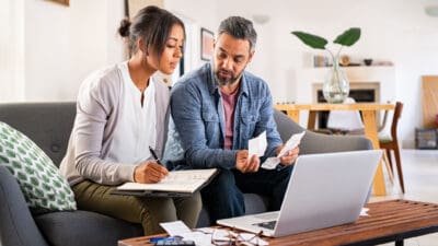 A couple calculate their budget and finances at home using laptop and calculator.