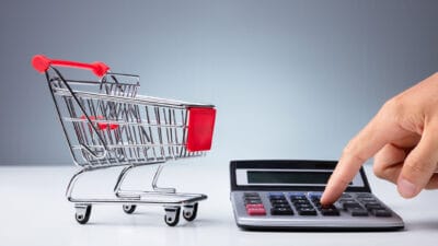 Close-up Of Empty Shopping Cart Near Person's Hand Using Calculator Over White Desk