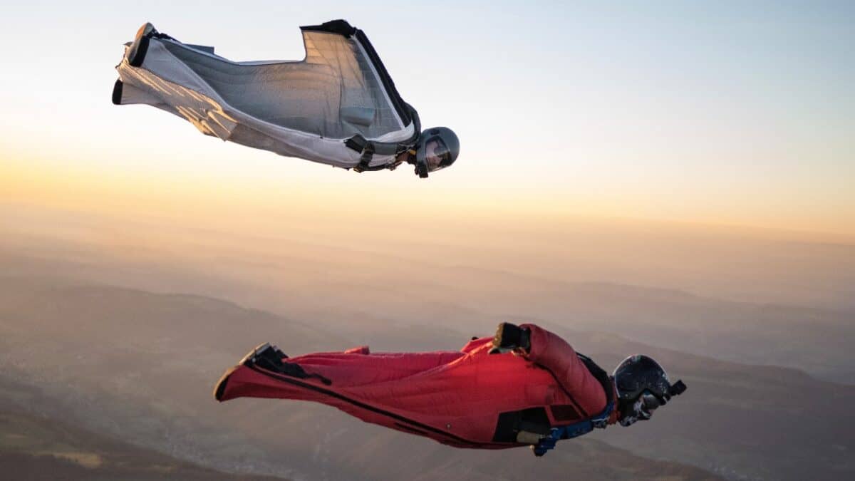 Two people in flying suits and helmets cruise in mid-air high above the earth with arms outstretched and the sun on the horizon.