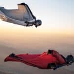 Two people in flying suits and helmets cruise in mid-air high above the earth with arms outstretched and the sun on the horizon.