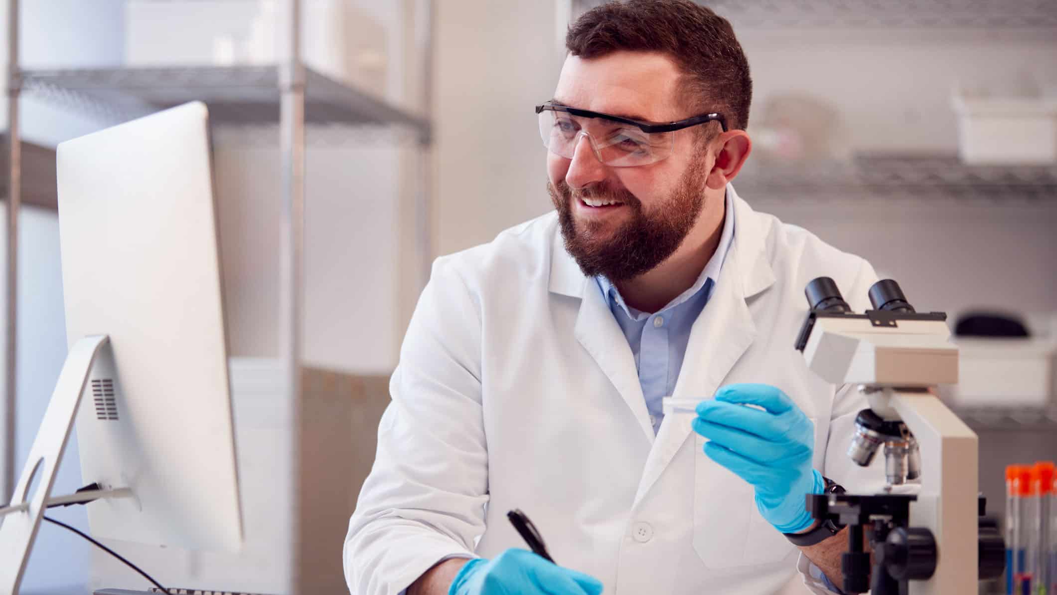 Male Lab Worker Wearing White Coat Recording Test Results On Computer.