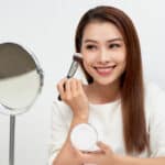 Happy woman looking in the mirror and applying cosmetic with a big brush.