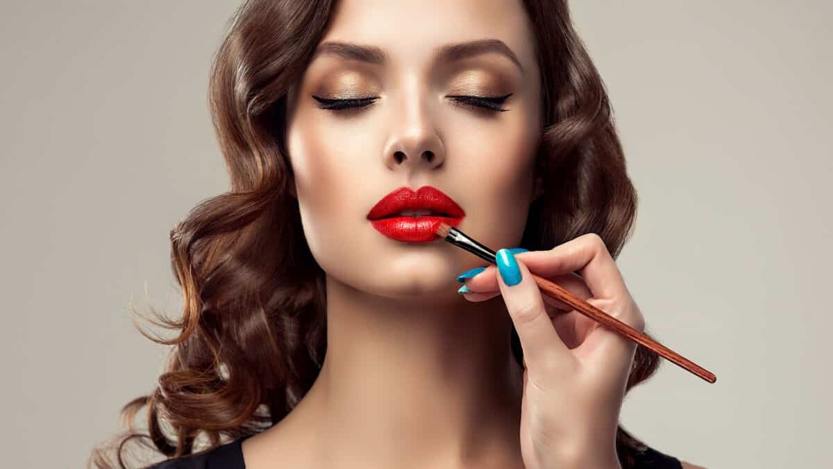 Graceful hands of professional make-up master, with the blue manicure on the nails, is painting in the red colour lips of splendid young woman.