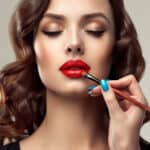 Graceful hands of professional make-up master, with the blue manicure on the nails, is painting in the red colour lips of splendid young woman.