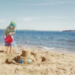 A young boy plays on a sunny beach pouring water from a bucket into a moat he has built around a sandcastle that is decorated with colourful shells.