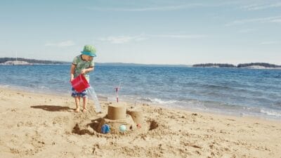 A young boy plays on a sunny beach pouring water from a bucket into a moat he has built around a sandcastle that is decorated with colourful shells.