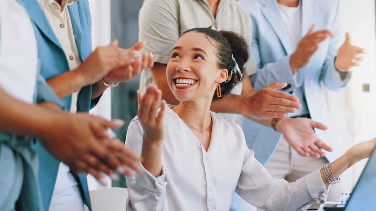 A young office worker is surrounded by peers who are clapping and congratulating her.