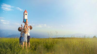 Three small children reach up to hold a toy rocket high above their heads in a green field with a blue sky above them.