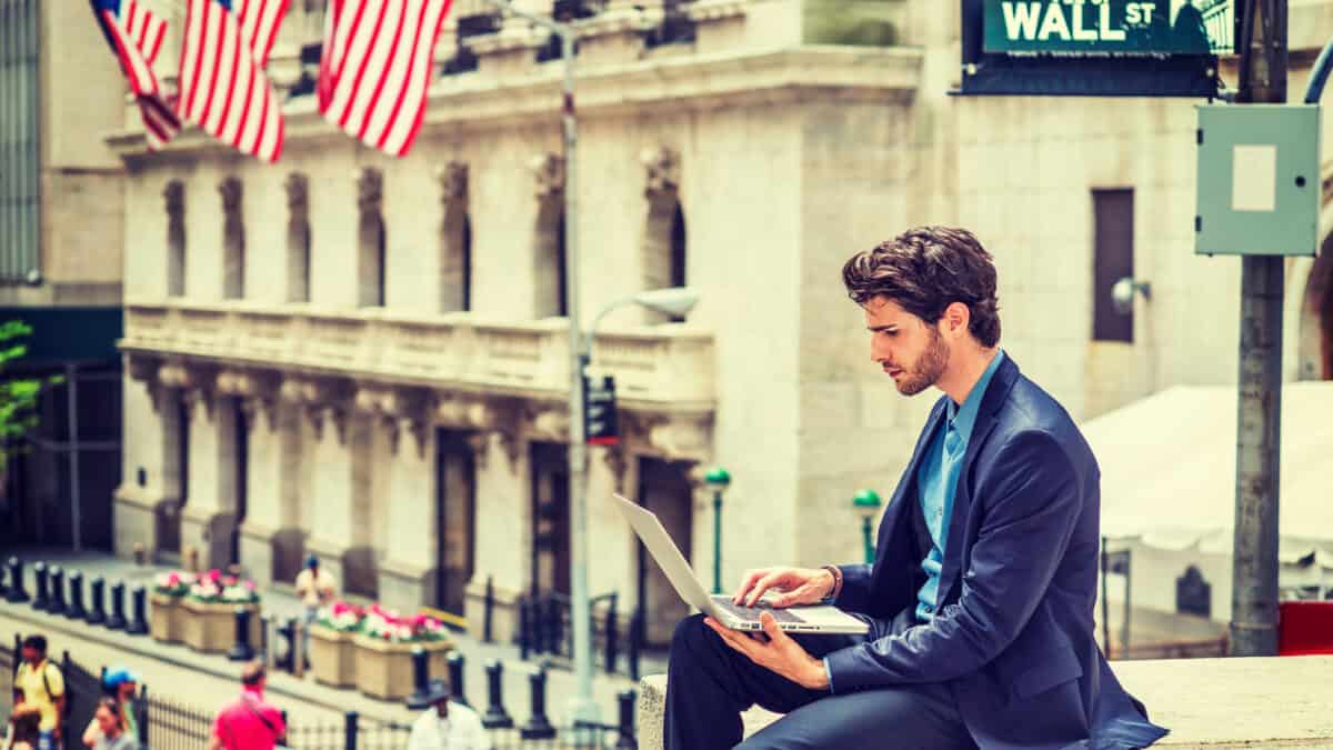 Businessman working on street in New York. Dressing in blue suit, a young guy with beard, sitting outside office building, looking down, reading, typing on laptop computer.