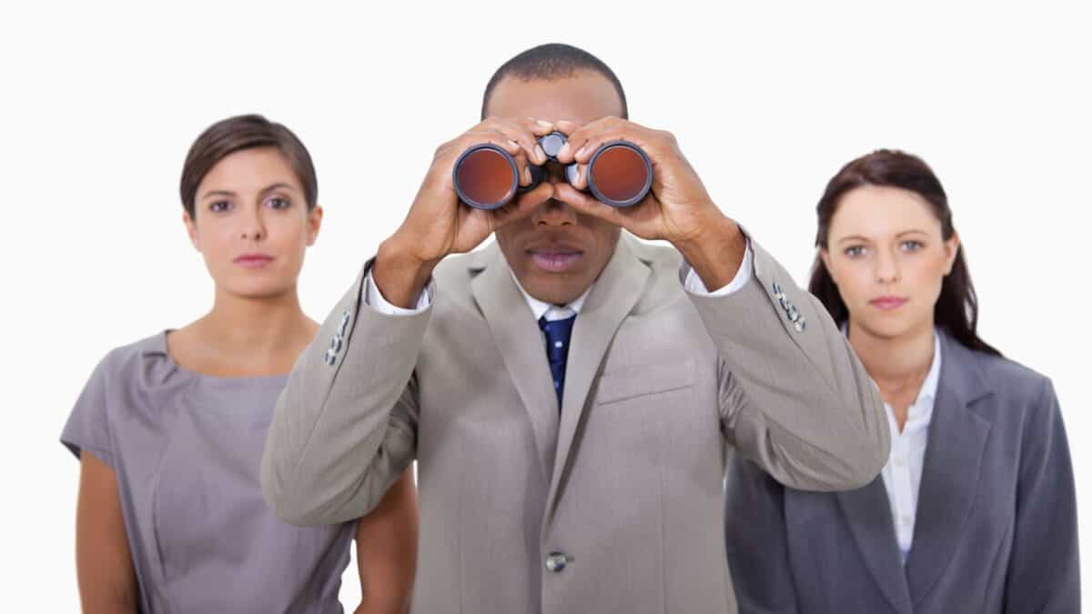 A man in a business suit peers through binoculars as two businesswomen stand beside him looking straight ahead at the camera.