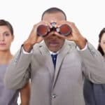 A man in a business suit peers through binoculars as two businesswomen stand beside him looking straight ahead at the camera.