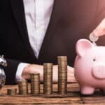 A businessman in a suit adds a coin to a pink piggy bank sitting on his desk next to a pile of coins and a clock, indicating the power of compound interest over time.