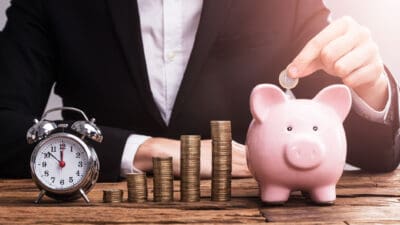 A businessman in a suit adds a coin to a pink piggy bank sitting on his desk next to a pile of coins and a clock, indicating the power of compound interest over time.