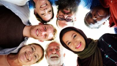 Seven men and women of different ages and nationalities put their heads together and smile as they look down at the camera.
