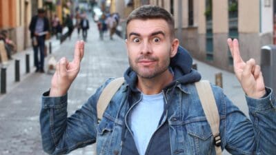 A young man wearing a backpack in a city street crosses his fingers and hopes for the best.