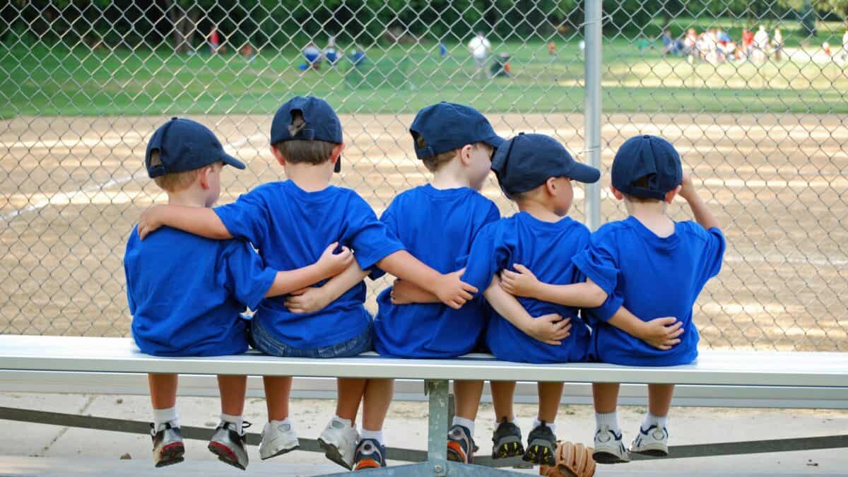 Five young boys wearing small caps sit on a bench together watching a baseball game.
