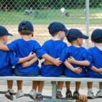 Five young boys wearing small caps sit on a bench together watching a baseball game.