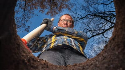 View from below of a man with a shovel standing by a hole he has dug in the garden, with blue sky in the background.